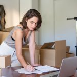 Empreendedora organiza seu negócio de roupas em um escritório doméstico, diante de um notebook e caixas de produtos.