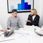 Profissionais reunidos em torno de uma mesa discutem dados e estratégias com laptops e gráficos.