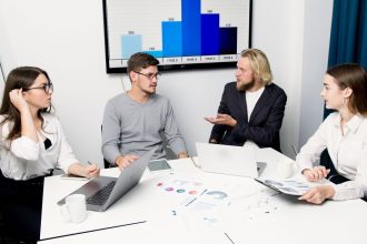 Profissionais reunidos em torno de uma mesa discutem dados e estratégias com laptops e gráficos.