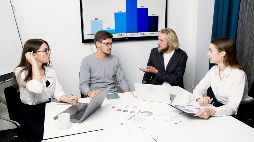 Profissionais reunidos em torno de uma mesa discutem dados e estratégias com laptops e gráficos.