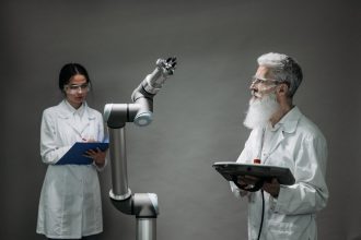 Dois pesquisadores trabalham com um braço robótico em um laboratório, representando pesquisa, inovação e desenvolvimento tecnológico.